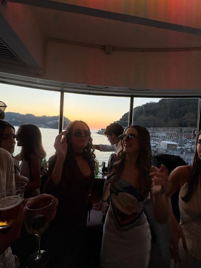 Chicas con gafas de sol bailan junto a los ventanales de una sala, con el mar y un cielo naranja de atardecer detrás.
