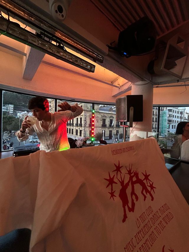 Un DJ con camisa blanca sostiene una copa en la cabina bajo luz anaranjada; en primer plano, una camiseta con estampado rojo de La Nuit Club.