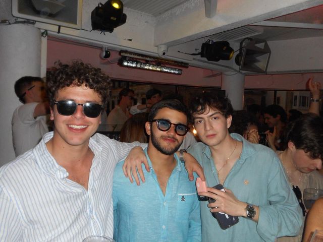 Tres chicos posan abrazados en un local lleno de gente, con focos y altavoces en el techo; dos de ellos llevan gafas de sol.