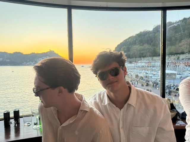 Dos chicos con camisas claras y gafas de sol posan junto a un ventanal, con la bahía y un cielo naranja de atardecer detrás.