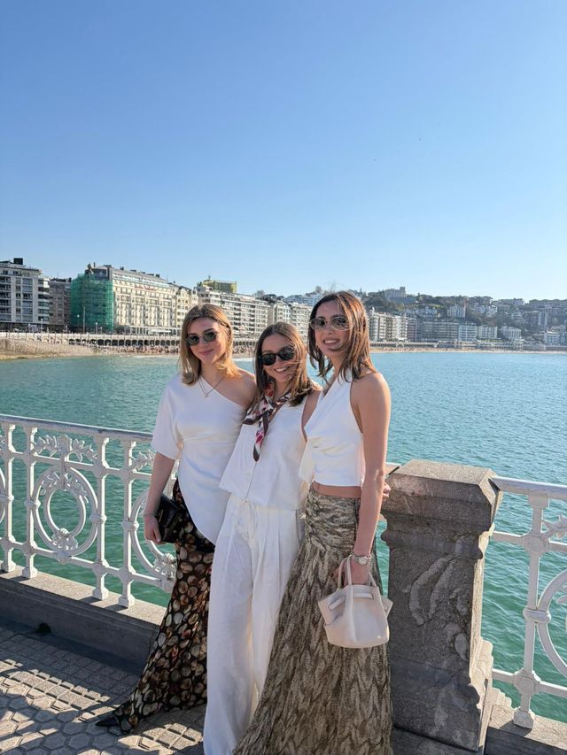 Tres amigas con gafas de sol posan junto a una barandilla blanca ornamentada, con el mar turquesa y la ciudad detrás en un día soleado.