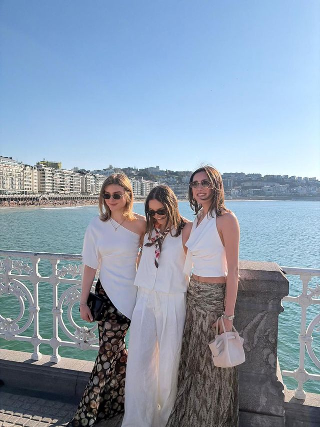 Tres amigas con gafas de sol posan muy juntas frente a la bahía, junto a una barandilla blanca, con el cielo azul y la playa al fondo.