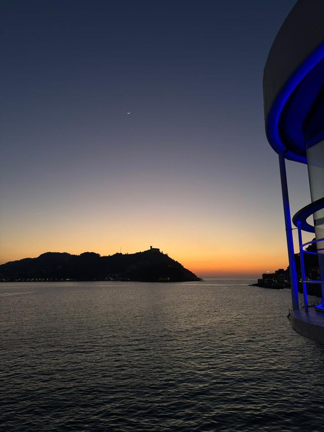 El cielo del atardecer pasa del azul al naranja sobre la bahía, con una colina en silueta, una luna creciente y un edificio curvo con luces azules.