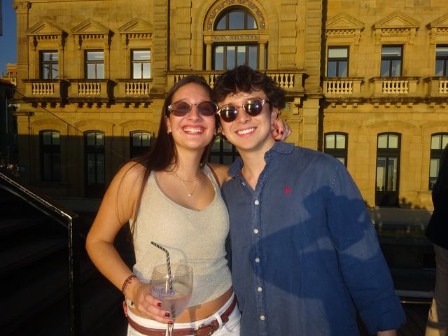 Una chica y un chico con gafas de sol ríen abrazados frente a un edificio señorial bañado por la luz dorada del atardecer.