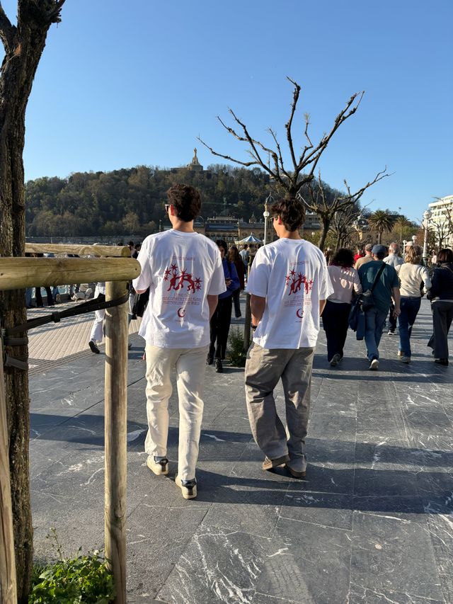 Dos chicos con camisetas blancas de estampado rojo caminan de espaldas por un paseo soleado, entre la gente y con una colina al fondo.