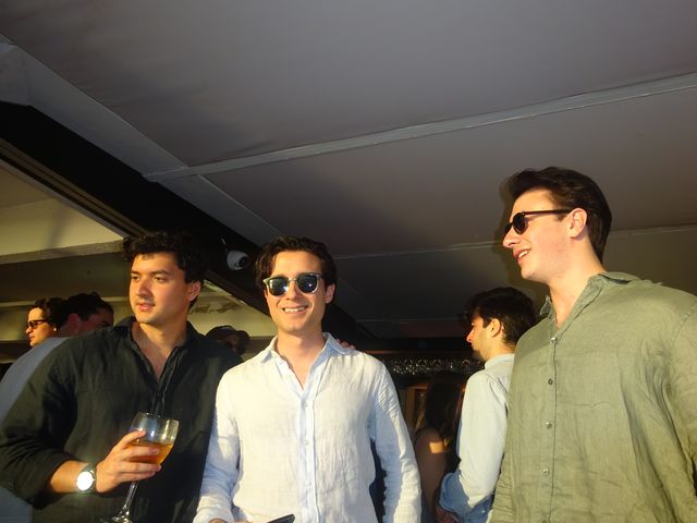 Tres chicos charlan bajo un techo bajo en el interior del local, dos con gafas de sol y uno con una copa en la mano.