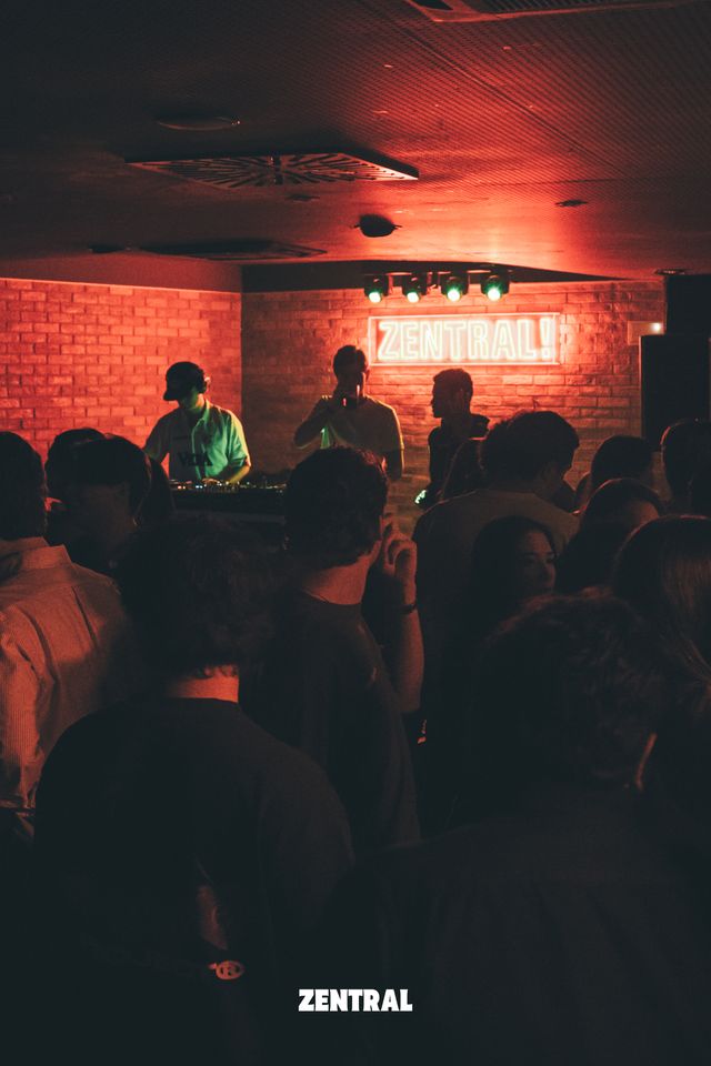 Un DJ con gorra mezcla bajo el letrero de neón Zentral, en una sala de ladrillo con luz roja y el público de espaldas en primer plano.