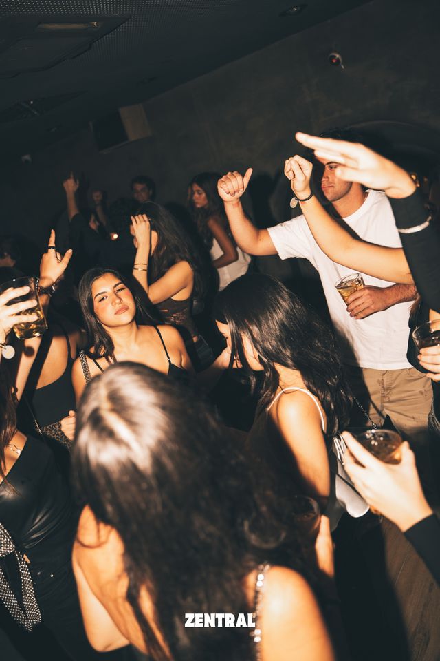 Un grupo de chicas y chicos baila con los brazos en alto y vasos en la mano, en una sala oscura de paredes grises.