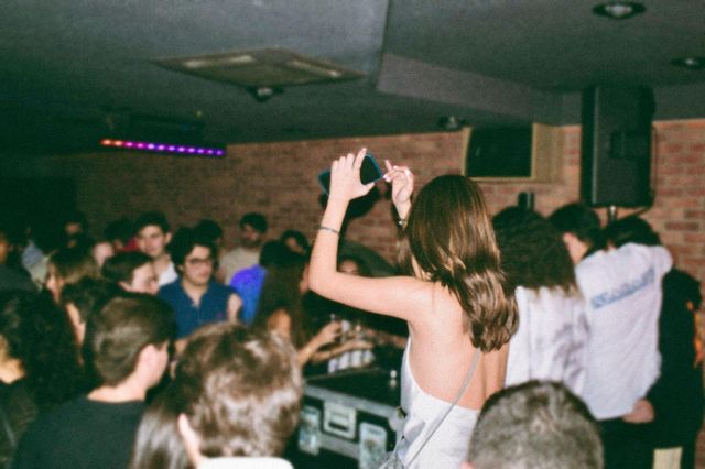 Una chica de espaldas sostiene el móvil en alto junto a la cabina del DJ, con la sala llena y la pared de ladrillo detrás.