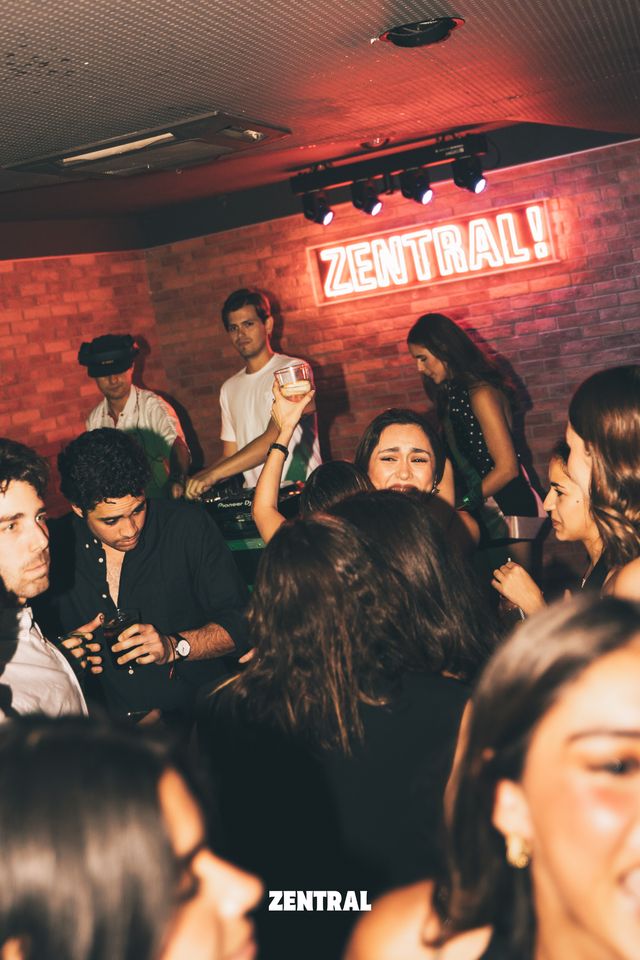 Dos chicos pinchan tras la mesa de mezclas bajo el neón rojo de Zentral, con la pared de ladrillo detrás y el público delante.