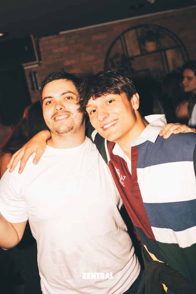 Dos chicos posan abrazados y sonriendo a la cámara, uno con camiseta blanca y otro con polo de rayas, con una pared de ladrillo detrás.