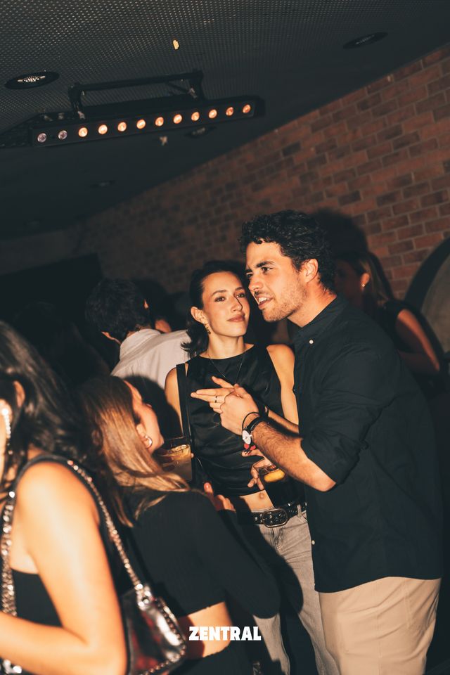 Un chico de camisa negra y una chica con top negro conversan muy cerca, con una barra de luces cálidas en el techo y una pared de ladrillo detrás.