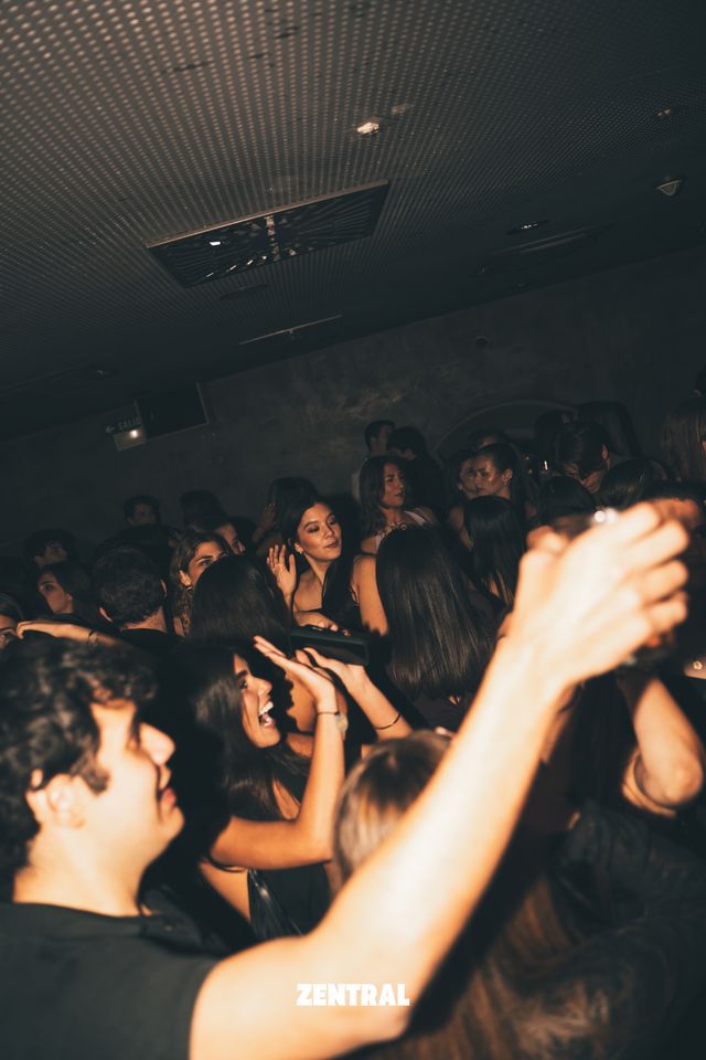 Chicos y chicas bailan apretados en una sala oscura, con las manos en alto y el brazo de un chico cruzando el primer plano.