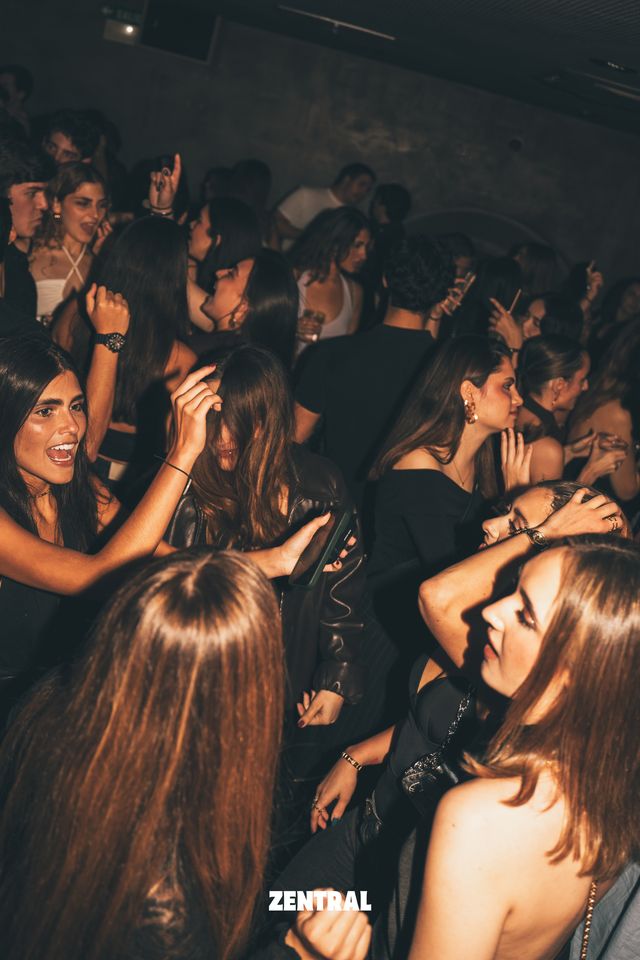 Un grupo de chicas baila y canta con el pelo suelto entre la multitud de una sala oscura, algunas con las manos en la cabeza.