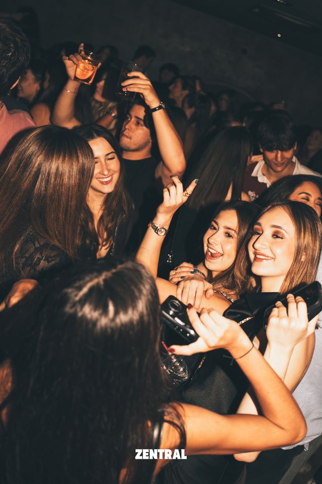 Un grupo de chicas sonríe a la cámara en plena fiesta, una de ellas sujetando una cámara compacta, con gente alzando sus copas detrás.