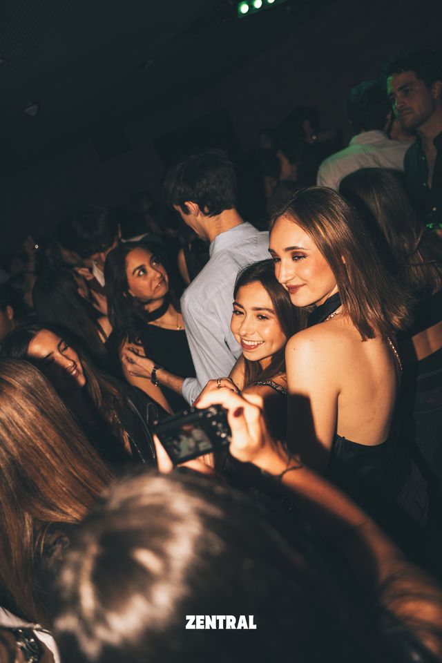 Dos chicas posan muy juntas y sonríen a la cámara entre la multitud, con un chico de camisa celeste detrás y una cámara en primer plano.
