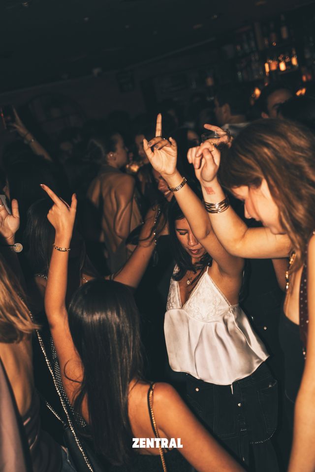 Varias chicas bailan con los brazos en alto bajo una luz cálida, una de ellas con top blanco y los ojos cerrados, y botellas al fondo.