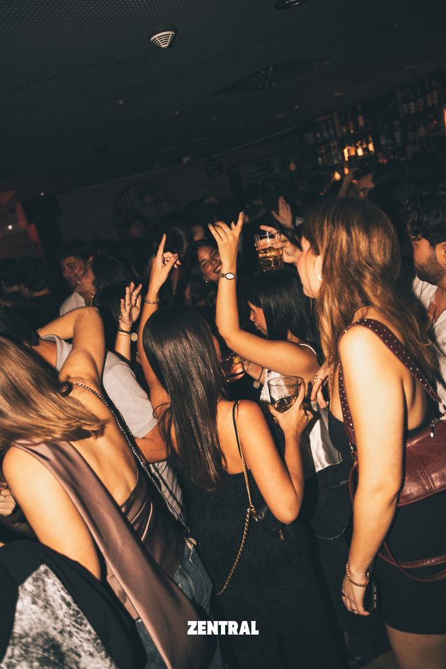 Un grupo de chicas baila de espaldas con los brazos en alto y copas en la mano, con una estantería de botellas iluminada al fondo.