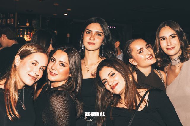Seis chicas posan muy juntas y sonríen a cámara en un bar de luz tenue, con botellas iluminadas al fondo.