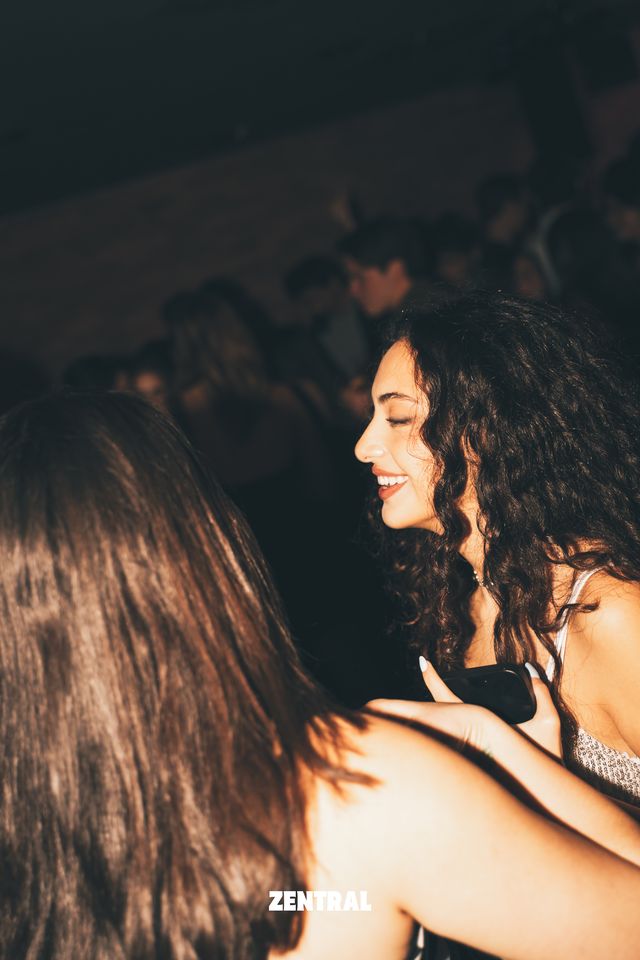 Una chica de pelo largo y rizado sonríe de perfil en una sala oscura, con el pelo de otra chica de espaldas ocupando el primer plano.
