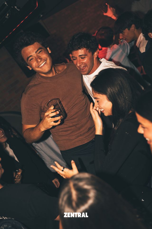 Dos chicos y una chica ríen juntos bajo una luz cálida; el de la camiseta marrón sostiene un vaso, con una pared de ladrillo al fondo.