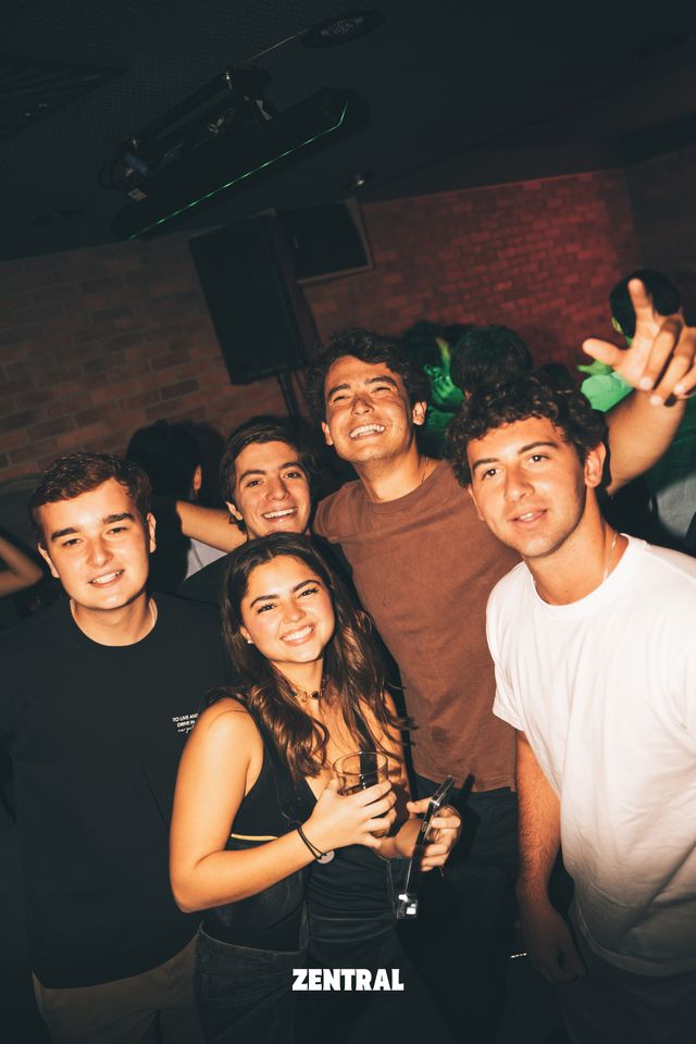 Un grupo de chicos y una chica posa sonriendo a la cámara, con una pared de ladrillo, un altavoz y luz verde detrás.