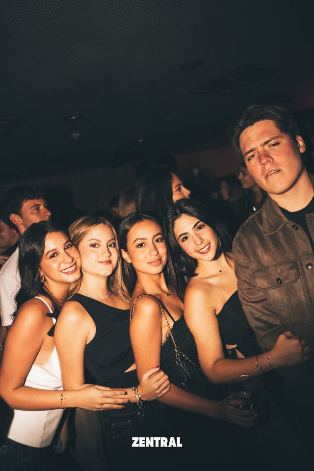 Cuatro chicas posan abrazadas y sonrientes junto a un chico serio con chaqueta marrón, en una sala oscura con más gente al fondo.