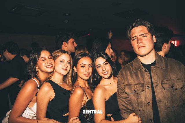 Cuatro chicas posan sonrientes, una con los ojos cerrados, junto a un chico serio de chaqueta marrón, con más gente al fondo.