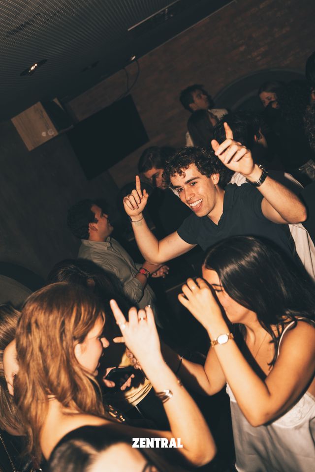 Un chico de pelo rizado sonríe y baila con los brazos en alto tras dos chicas, en una sala oscura de paredes de ladrillo.