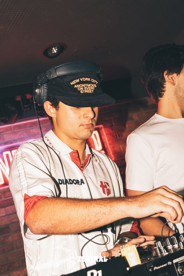 Un DJ con gorra negra, auriculares y camiseta blanca de cuello rojo mezcla en los platos; a su lado, un chico de blanco.