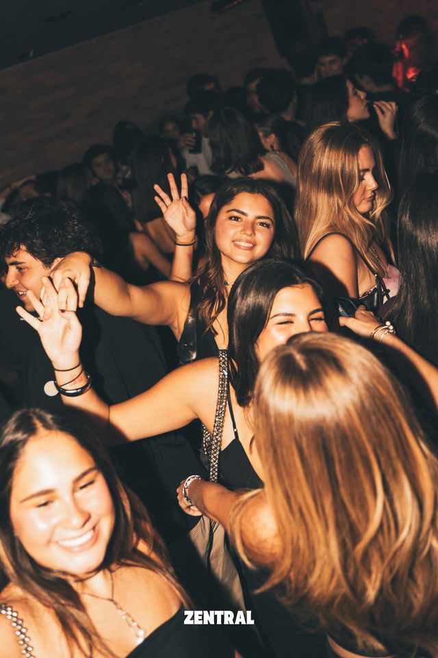 Un grupo de chicas sonríe y baila en una pista abarrotada, una de ellas con la mano en alto, con una pared de ladrillo al fondo.
