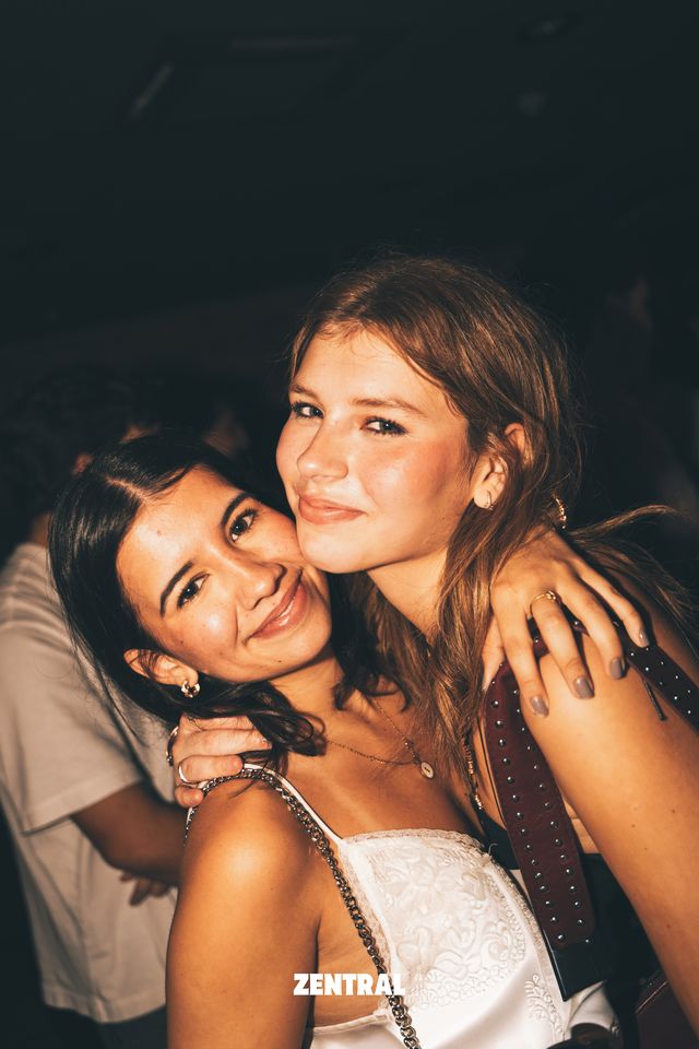 Dos chicas se abrazan mejilla con mejilla y sonríen a la cámara en una sala oscura, una con top blanco de encaje.