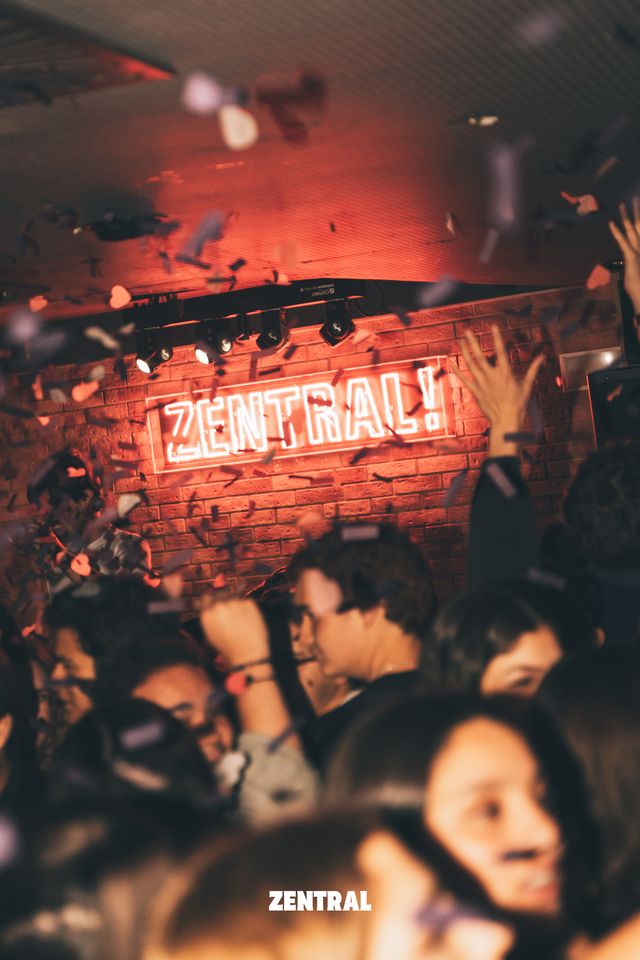 Un grupo baila entre confeti bajo el letrero de neón rojo de Zentral, con una pared de ladrillo detrás y una mano alzada.