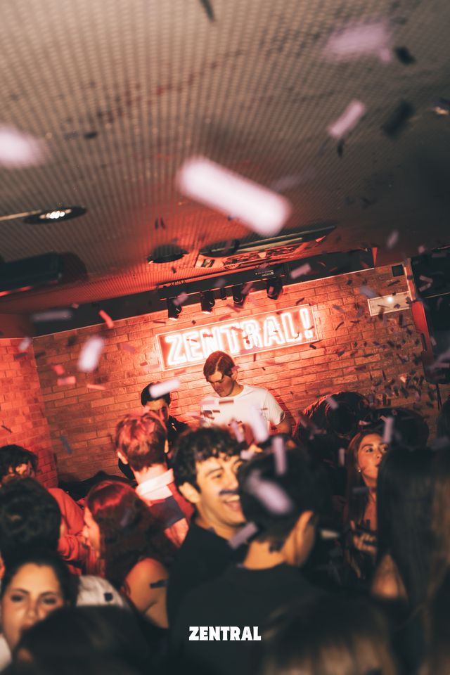Vista general de la sala con el neón rojo de Zentral sobre la pared de ladrillo, gente sonriendo en primer plano y confeti en el aire.