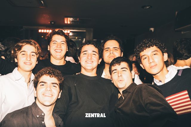 Un grupo de chicos posa muy apretado y sonríe a la cámara en una sala oscura, con uno de ellos haciendo morritos.