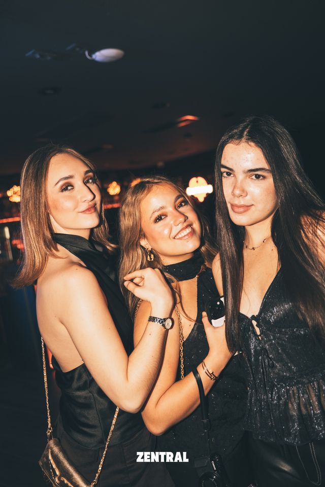 Tres amigas vestidas de negro posan juntas ante la cámara en un local oscuro con luces cálidas al fondo.