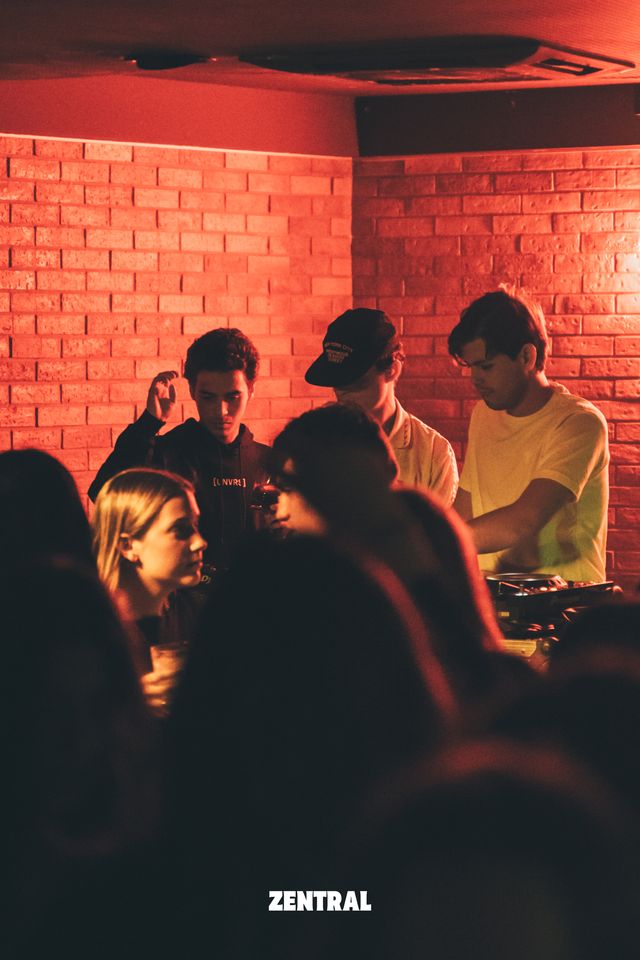 Tres chicos en la cabina de DJ bajo una intensa luz roja, uno de ellos mezclando; delante, una chica con una copa y el público a oscuras.