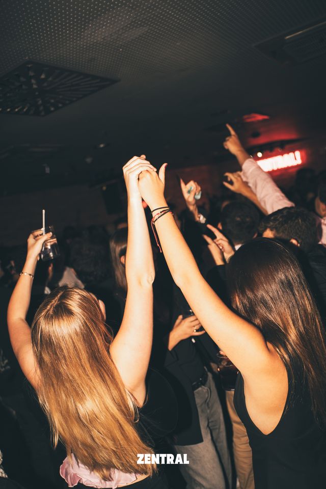 Dos chicas de espaldas levantan los brazos con las manos entrelazadas, en una sala oscura con un cartel de neón rojo al fondo.