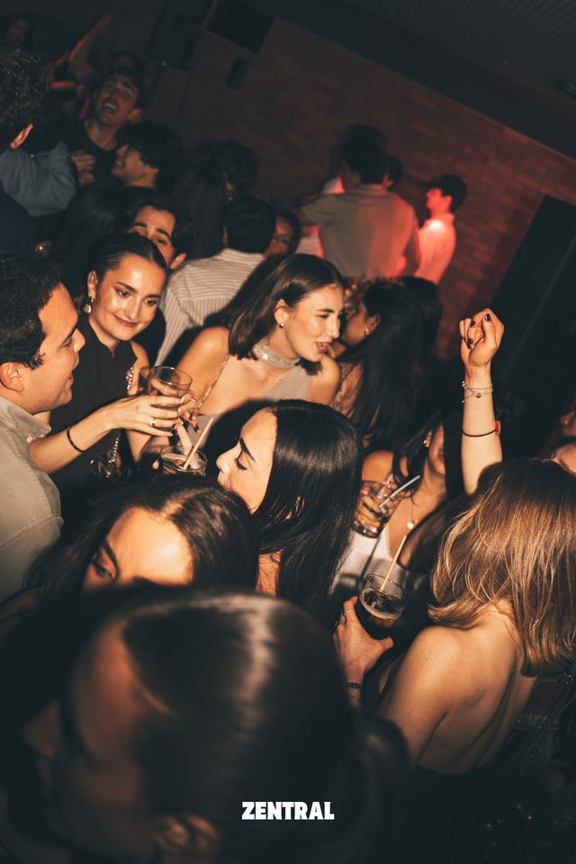 Chicas y chicos bailan muy juntos con copas y pajitas en la mano, delante de una pared de ladrillo con luz rojiza.