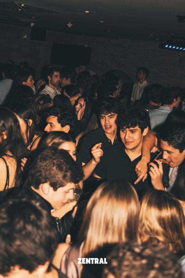Un grupo de chicos y chicas se apretuja en la pista bajo luces tenues; en el centro, dos chicos de negro miran a cámara.