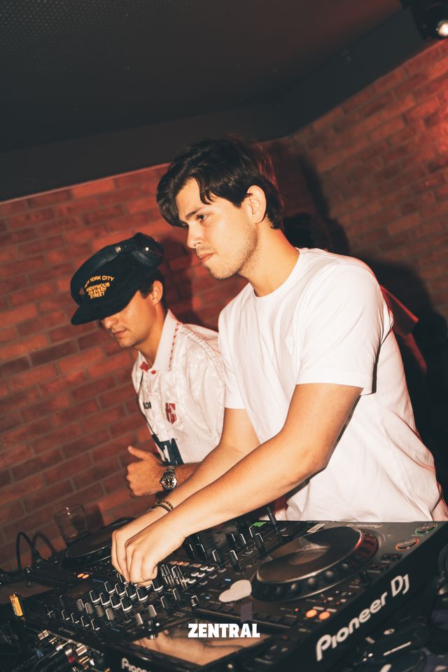Dos DJ mezclan juntos en los platos, uno con camiseta blanca y otro con gorra negra y auriculares, ante una pared de ladrillo rojo.