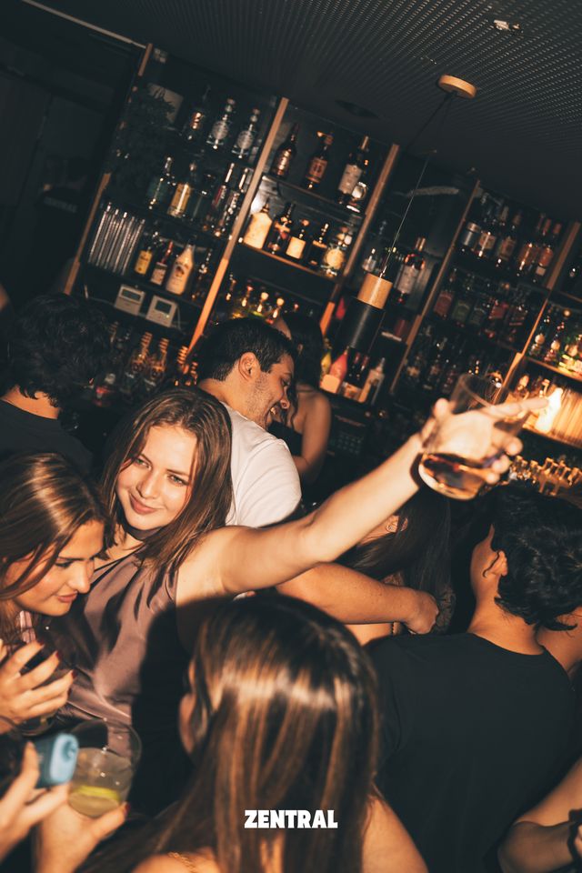Una chica sonríe a cámara con el brazo estirado y una copa en la mano, junto a un chico que ríe, ante estantes llenos de botellas.