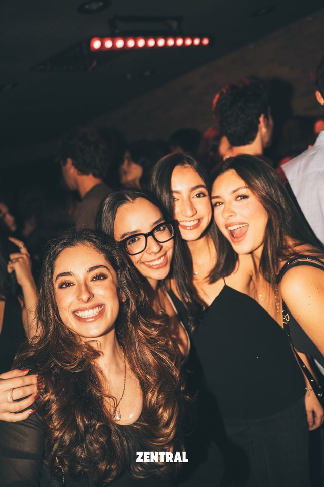 Cuatro amigas posan juntas y sonrientes ante la cámara, una de ellas con gafas, con una tira de luces rojas en el techo.
