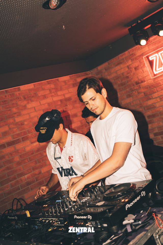 Dos DJ mezclan en la cabina, uno con camiseta blanca y otro con gorra negra, junto a una pared de ladrillo con un cartel de neón.