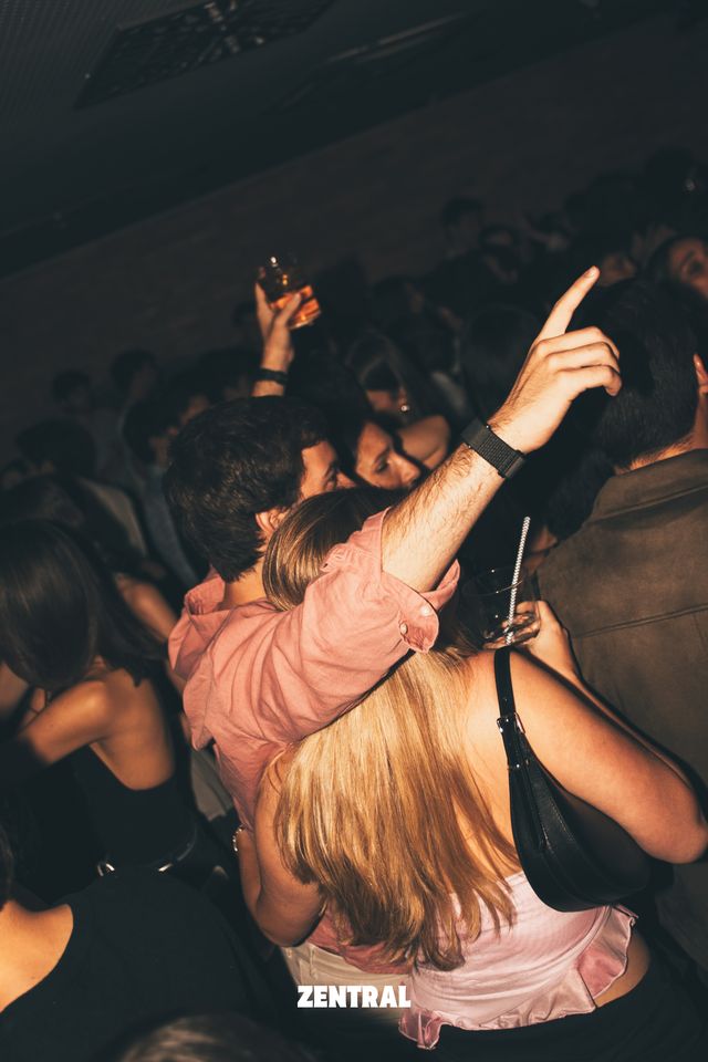 Un chico con camisa rosa levanta el brazo señalando hacia arriba mientras baila junto a una chica de espaldas con melena rubia.