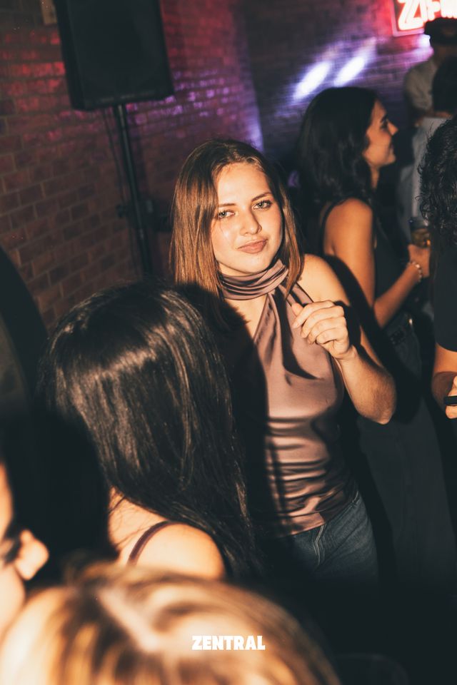 Una chica de pelo castaño y top satinado mira a cámara con media sonrisa, junto a un altavoz y una pared de ladrillo con luz rosada.