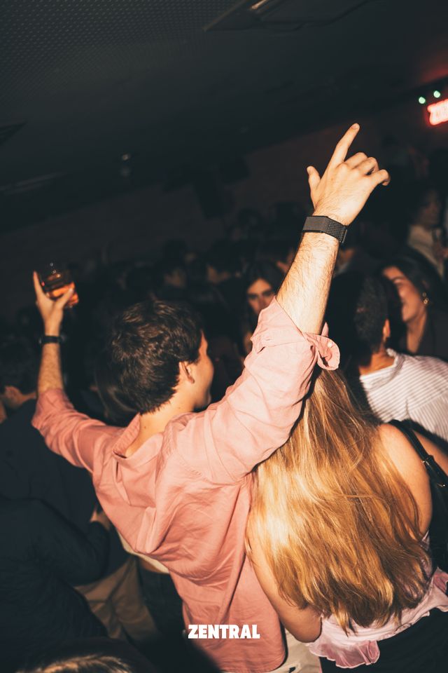 Un chico con camisa rosa baila con un brazo en alto y una copa en la otra mano, junto a una chica de larga melena rubia, entre la multitud.