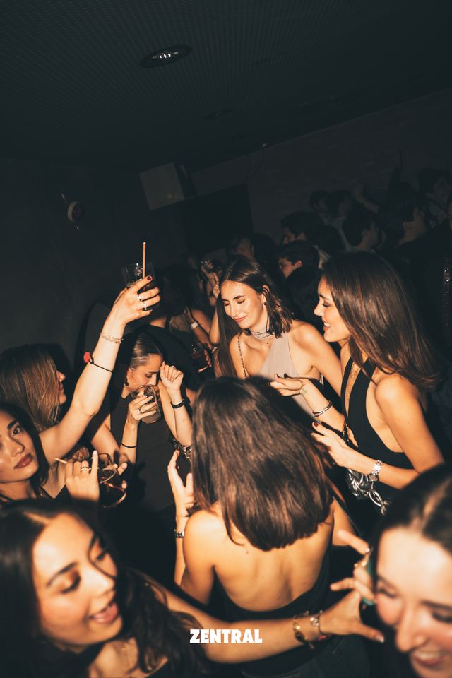 Un grupo de chicas baila muy junto en la pista oscura, sonriendo y con copas en la mano, una de ellas con el brazo en alto.