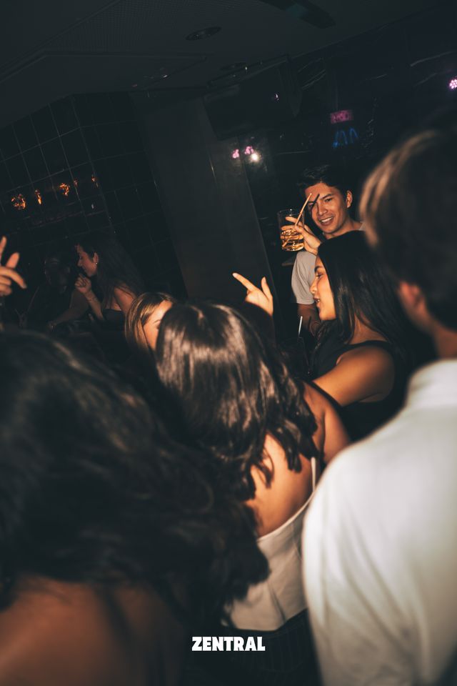 Un chico sonríe con una copa en la mano junto a una chica de perfil, al fondo de la sala, detrás de varias personas de espaldas.