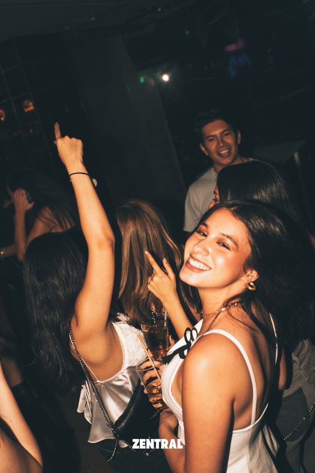 Una chica con top blanco sonríe a cámara mirando por encima del hombro, mientras otra baila con el brazo en alto y un chico ríe detrás.